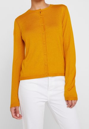 Frau trägt eine senfgelbe, langärmelige Strickjacke mit Knöpfen, kombiniert mit weißen Hosen, gezeigt vom Hals bis zur Hüfte.