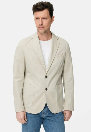 Mężczyzna w beżowej marynarce na dwa guziki, białej koszuli i niebieskich dżinsach, stojący i patrzący na bok na jednolitym tle.