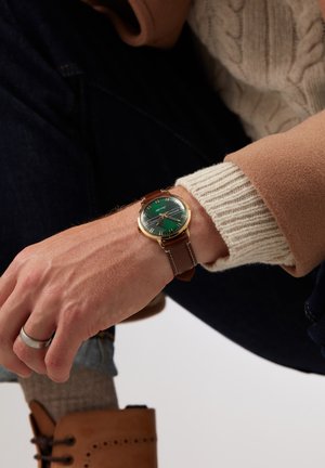 Montre-bracelet en cuir marron avec un cadran vert, un boîtier en or et des marqueurs d'heure en argent. Portée sur une main, avec un pull en tricot et un jean visibles.