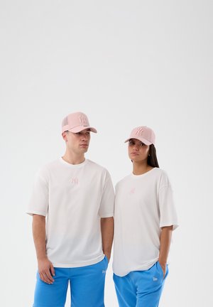 Due persone che indossano magliette bianche abbinate con loghi rosa, cappellini da baseball rosa e pantaloncini blu brillanti, in piedi davanti a uno sfondo bianco uniforme.
