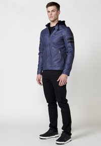 Chaqueta de cuero azul con cremallera frontal, capucha y dos bolsillos en el pecho con cremallera; cuenta con acentos en rayas negras en las mangas; combinada con pantalones negros.