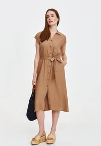 Beżowa sukienka z guzikami, związywana w talii, z krótkimi rękawami i bocznymi kieszeniami. Noszona z plecionymi sandałami i czarną torebką na szydełku.