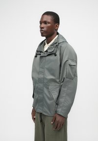 Γκρίζα φούτερ ζακέτα με φερμουάρ μπροστά, ρυθμιζόμενο κορδόνι και μεγάλες πλαϊνές τσέπες. Κατασκευασμένη από λείο, ελαφρύ ύφασμα. Λεπτομέρεια με τσέπη μπροστά.