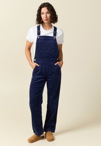 Marineblaue Corduroy-Overalls mit Schulterträgern, einer Fronttasche, seitlichen Taschen und lockerer Passform, kombiniert mit einem weißen T-Shirt und braunen Clogs.
