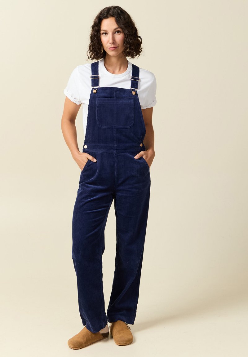 Marineblaue Corduroy-Overalls mit Schulterträgern, einer Fronttasche, seitlichen Taschen und lockerer Passform, kombiniert mit einem weißen T-Shirt und braunen Clogs.