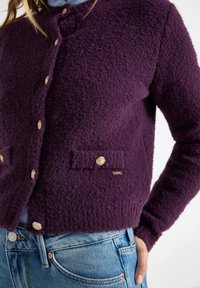 Purple wool cardigan with a textured finish, featuring rounded gold buttons and two front pockets, paired with a blue shirt.