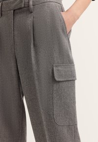Grau melierte Wollmischungshosen mit strukturierter Oberfläche, die seitliche Taschen und eine Klapptasche am Bein für zusätzliche Funktionalität aufweisen.