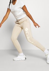 Beige sweatpants med en vit "Columbia" -logotyp; elastisk midja, sidofickor och ribbade muddar. Kombineras med en ljusgrå t-shirt och vita sneakers.