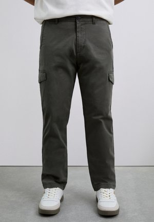 Persona con pantalones cargo verde oscuro y zapatillas blancas de pie sobre un suelo gris frente a una pared lisa.