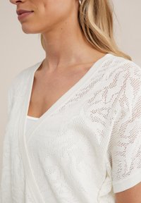 Cardigan tricoté blanc avec un col en V et des manches courtes, présentant un motif texturé et un design à mailles ouvertes.