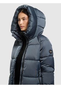 Jakna punjena perjem u tamnosivoj boji, s velikom kapuljačom, prošivenim uzorkom i zatvaračem. Na lijevom rukavu nalazi se zakrpa s logom.