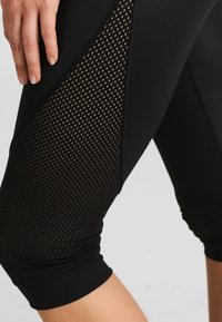 Zbliżenie na czarne legginsy sportowe z siateczkowymi panelami po bokach nóg, na których widać rękę spoczywającą na udzie.