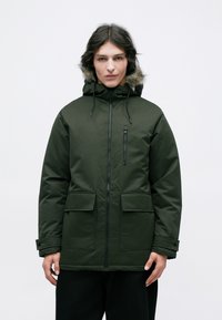 Grøn isoleret parka-jakke med en hætte foret med pels, frontlynlås, to store forlommer og en lille brystlomme. Glat, slidstærkt stof.