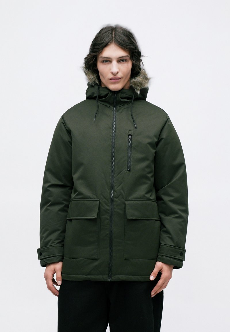 Grøn isoleret parka-jakke med en hætte foret med pels, frontlynlås, to store forlommer og en lille brystlomme. Glat, slidstærkt stof.