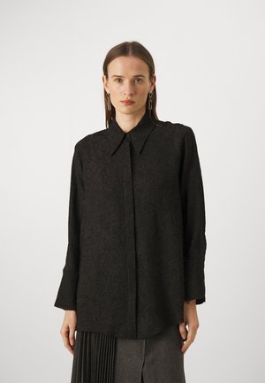 Camicia nera con bottoni in tessuto strutturato, dotata di colletto, maniche lunghe e intricati motivi floreali. Abbinata a una gonna plissettata.
