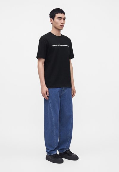 Νέος άνδρας που φοράει μαύρο T-shirt Emporio Armani, άνετο μπλε τζιν και μαύρα αθλητικά παπούτσια μπροστά σε απλό φωτεινό φόντο.