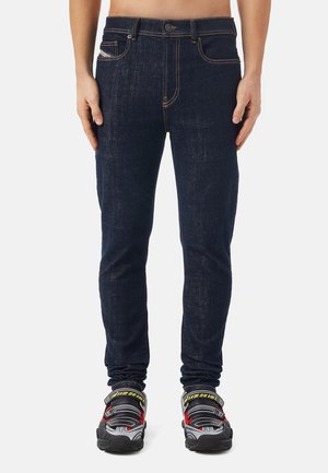 Homme debout, portant un jean slim bleu foncé et des baskets gris, noir, rouge et jaune sur un fond uni.