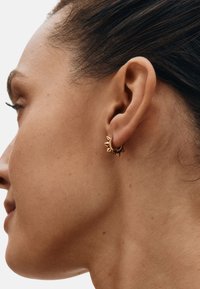 Guldcreol ørering med spidse detaljer og en tekstureret overflade. Båret i øret og supplerer en neutral hudtone. Minimalistisk design.