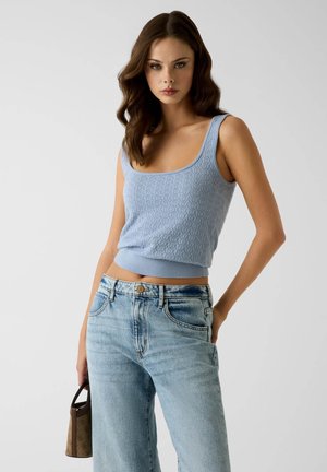 Jeune femme aux cheveux bruns ondulés portant un débardeur en tricot bleu clair, un jean bleu délavé et tenant un petit sac à main marron.