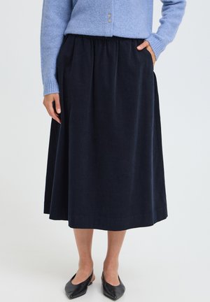 Femme portant une jupe midi bleu marine avec poches, un pull bleu clair et des ballerines slingback noires à bout pointu, debout devant un fond uni.