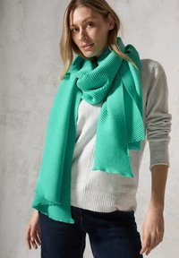 Turkis ribbet tørklæde viklet omkring halsen, med blød tekstur og frynser i kanterne, parret med en lys grå sweater.