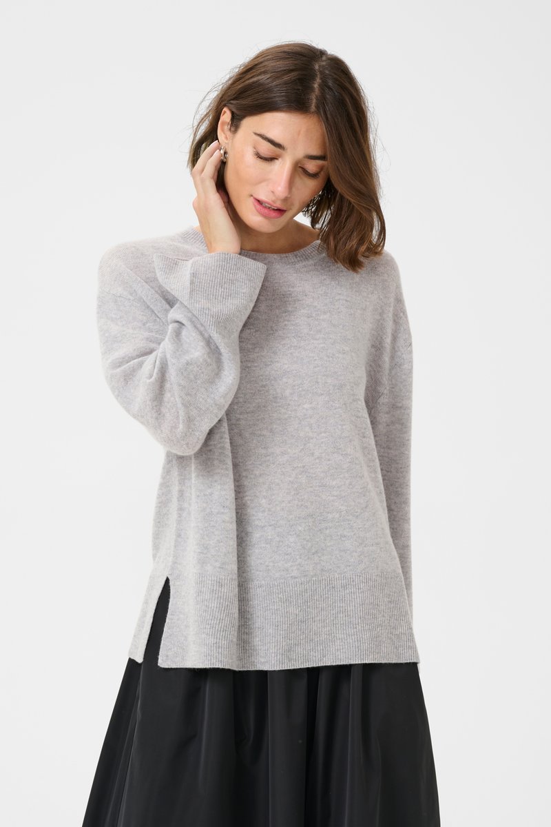Svetlo siva pletena pulover s okroglim ovratnikom, stranskimi razporki in rebrastimi manšeti. Mehka tekstura in sproščen kroj, kombiniran z črno krilom.