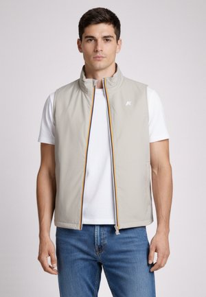 Giovane uomo che indossa un gilet beige senza maniche con cerniera sopra una maglietta bianca e jeans blu, in piedi davanti a uno sfondo semplice.