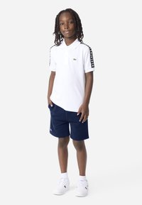 Wit poloshirt met korte mouwen, groen geborduurd logo en zwarte patroonrand. Gecombineerd met marineblauwe korte broek en witte sneakers.