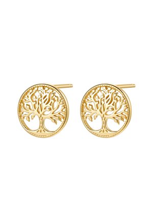 Woman wearing small round gold stud earrings with a tree design, touching her shoulder, dark hair and light-colored shirt visible.