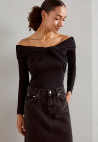 Svart off-shoulder långärmad topp med rynkad textil vid bysten, i kombination med en högmidjad svart denimkjol med knappstängning.