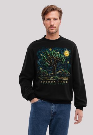 Mann in schwarzem Sweatshirt mit buntem Design des Joshua-Tree-Nationalparks und blauen Jeans, der mit einer Hand in der Tasche steht.