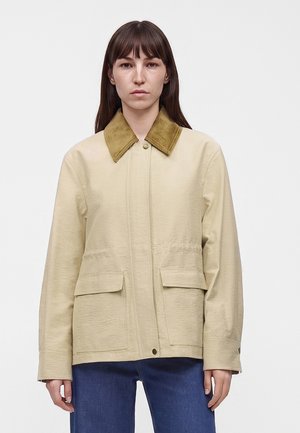 Femme aux cheveux bruns raides portant une veste beige avec un col en velours côtelé marron et un jean bleu, debout devant un fond uni.