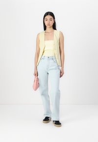 Gilet sans manches couleur crème sur un haut jaune à décolleté carré, associé à un jean bleu clair et des baskets noires. Tenant un sac à main rose.