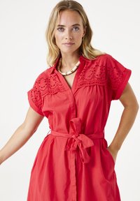 Rode korte jurk met decoratieve borduursels op de schouders, ceintuur in de taille, knoopsluiting aan de voorkant en zakken. Gladde katoenen stof.