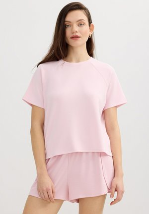 Mujer joven con cabello largo y castaño, vistiendo un top de manga corta rosa claro y pantalones cortos a juego, de pie frente a un fondo claro y liso.