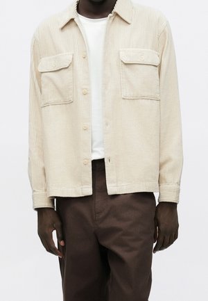 Mężczyzna w beżowej sztruksowej koszuli zapinanej na guziki z patkami na kieszeniach, na białej koszulce i brązowych spodniach, stojący na neutralnym tle.