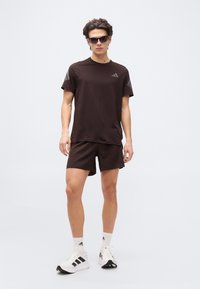 Νέος άνδρας που φοράει μαύρα αθλητικά ρούχα, λευκά παπούτσια Adidas, λευκές κάλτσες και σκούρα γυαλιά ηλίου, στέκεται μπροστά σε ένα απλό λευκό φόντο.