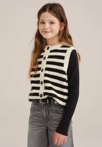 Černo-bílé pruhované pletené vestička s kulatým výstřihem a zapínáním na knoflík, nošená přes černou košili s dlouhým rukávem, spárovaná se šedými džíny.