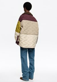Chaqueta acolchada en beige, burdeos y oliva. Presenta un cuello de piel marrón y mangas anchas. Combinada con unos vaqueros rectos azules.