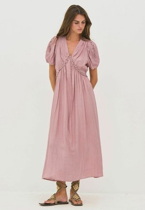 Kvinde iført en lilla ankellang kjole med puffede ærmer og flettede detaljer, kombineret med flade sandaler med guldbeslag, kigger ned.