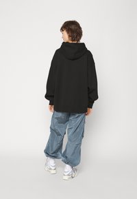 Sudadera negra de gran tamaño con capucha, combinada con pantalones cargo brillantes de color azul claro. El modelo lleva zapatillas blancas y está de pie con las manos a los lados.