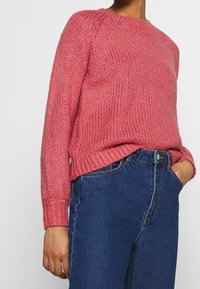 Pletený svetr z růžové texturované látky s kulatým výstřihem a žebrovanými manžetami, doplněný o modré džíny s vysokým pasem.