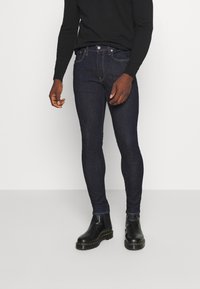 Jeans ajustados azul oscuro hechos de mezclilla, con cinco bolsillos, costuras en contraste y usados con botines negros. Fondo plano.