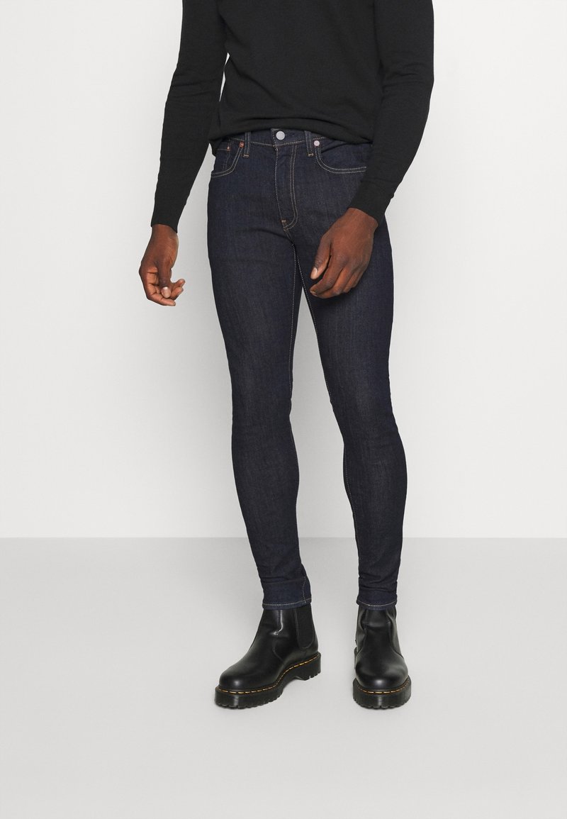 Jeans ajustados azul oscuro hechos de mezclilla, con cinco bolsillos, costuras en contraste y usados con botines negros. Fondo plano.