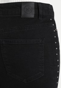 Schwarze Denim-Jeans mit strukturierten Oberflächen, ausgestattet mit einer Gesäßtasche und genieteten Akzenten an der Seite. Lederlogo-Patch am Bund.
