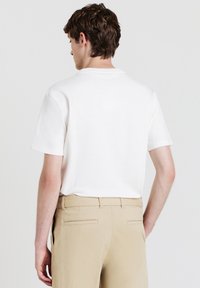 Uomo visto di spalle che indossa una camicia bianca a maniche corte con texture e pantaloni beige, in piedi davanti a uno sfondo chiaro e uniforme.