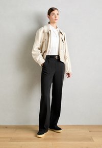 Beige übergroße Jacke mit vorderen Taschen, weißes Hemd, schwarze hochgeschnittene Hose und schwarze Schuhe mit gelben Sohlen auf einem Holzboden.