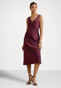 Bordó szatén ruha V-nyakkal, csipke részletekkel átlós varrások mentén, és térd alatti szegéllyel. Fekete, pántos magassarkú cipőkkel kiegészítve.