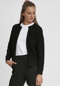Cardigan noir avec un motif en boules texturé, col et poignets côtelés, design à ouverture devant. Porté sur un t-shirt blanc à col ras du cou.