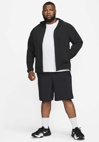 Zwarte jas met ritssluiting en capuchon, wit T-shirt met ronde hals, zwarte korte broek en zwarte sneakers met witte accenten. Casual sportkleding.